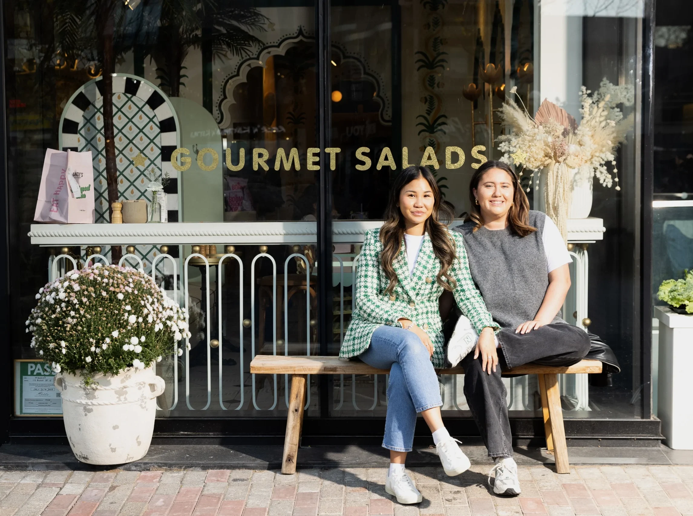 Two smiling women that manage Mandy's Salads sitting on bench outside storefront